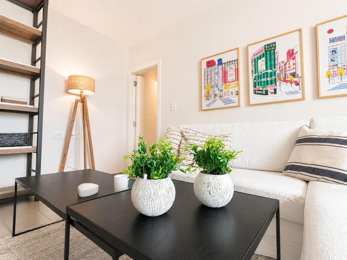 Sala luminosa con sofá blanco, mesas negras, plantas y cuadros coloridos en la pared.