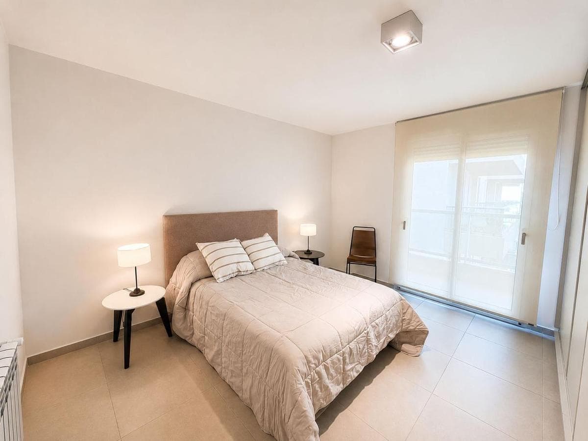 Dormitorio luminoso con cama beige, mesitas de noche con lámparas y un gran ventanal.