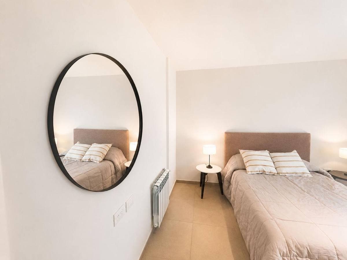 Dormitorio minimalista con un gran espejo ovalado de marco negro reflejando una cama acogedora.