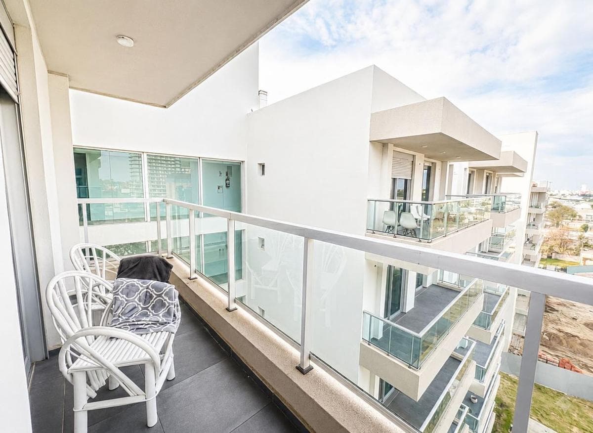 Balcón moderno con barandilla de vidrio, sillas blancas y vistas a edificios de apartamentos.